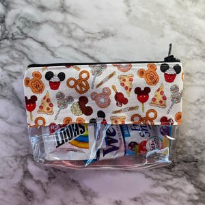 May include: A clear, rectangular cosmetic bag with a black zip and a patterned top. The top fabric features Mickey Mouse-themed food icons like pretzels, pizza, and cupcakes. The bag contains snacks.