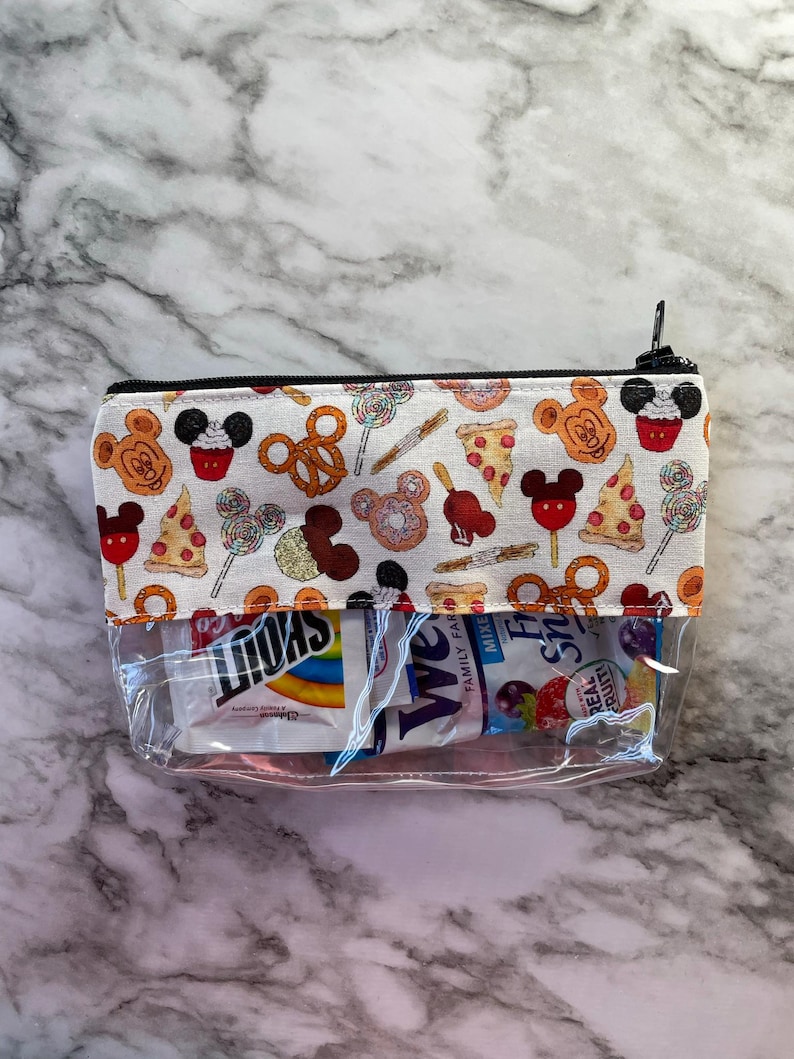 May include: A clear, rectangular cosmetic bag with a black zip and a patterned top. The top fabric features Mickey Mouse-themed food icons like pretzels, pizza, and cupcakes. The bag contains snacks.