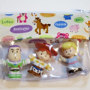 May include: A clear plastic zippered pouch with a white fabric lining featuring Toy Story characters and names like Lotso, Bullseye, Hamm, and Buzz. The pouch contains three small plastic figurines of Woody, Buzz Lightyear, and Jessie.