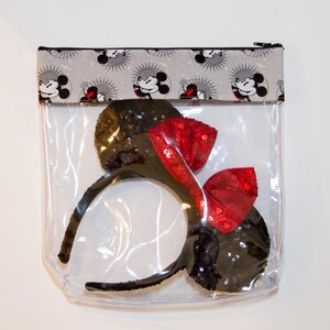 May include: A clear plastic zippered pouch with a black and white fabric top featuring a repeating pattern of Mickey Mouse. The pouch contains a pair of black mouse ears with a red bow.