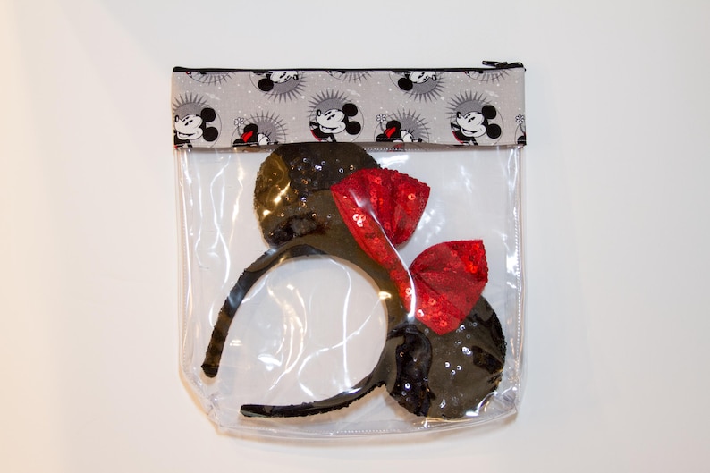 May include: A clear plastic zippered pouch with a black and white fabric top featuring a repeating pattern of Mickey Mouse. The pouch contains a pair of black mouse ears with a red bow.