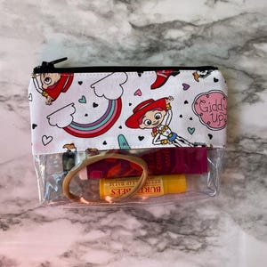 May include: A clear zippered pouch with a white background and a colorful pattern of rainbows, hearts, and cartoon characters. The pouch is open and contains a hair tie, a pink lip balm, and a yellow lip balm. The text "Giddy Up" is visible on the pouch.