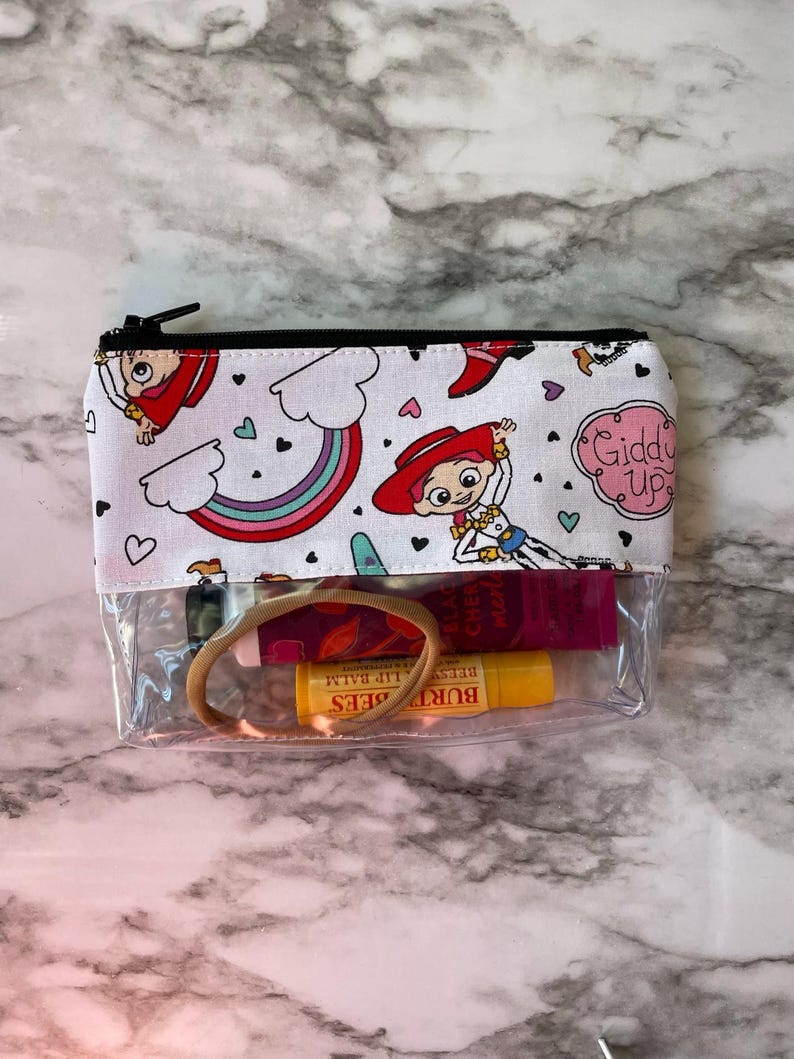 May include: A clear zippered pouch with a white background and a colorful pattern of rainbows, hearts, and cartoon characters. The pouch is open and contains a hair tie, a pink lip balm, and a yellow lip balm. The text "Giddy Up" is visible on the pouch.