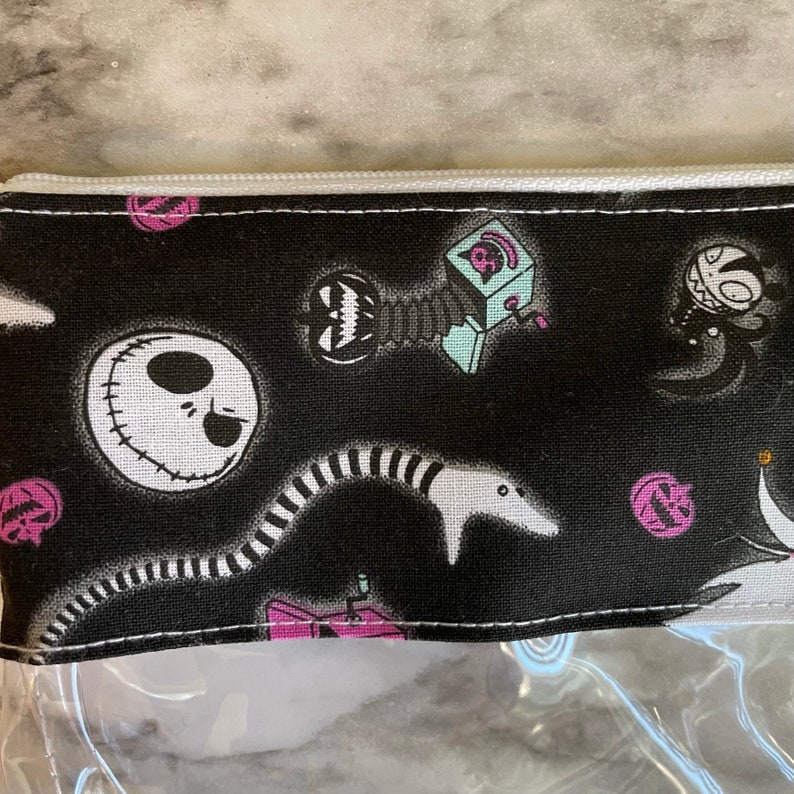 May include: Black zippered pouch with a clear plastic front. The pouch features a repeating pattern of characters from the Nightmare Before Christmas, including Jack Skellington, Oogie Boogie, and Sally.