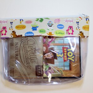 May include: A clear plastic zippered pouch with a Toy Story 3 DVD inside. The pouch has a fabric flap with a colorful pattern of Toy Story characters and their names. The flap reads "Jessie", "Woody", "Hamm", "Lotso", and "Buzz".