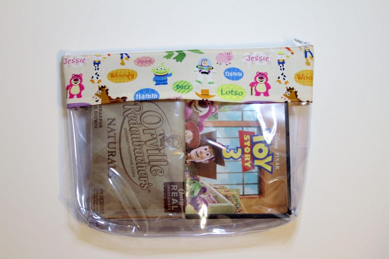 May include: A clear plastic zippered pouch with a Toy Story 3 DVD inside. The pouch has a fabric flap with a colorful pattern of Toy Story characters and their names. The flap reads "Jessie", "Woody", "Hamm", "Lotso", and "Buzz".