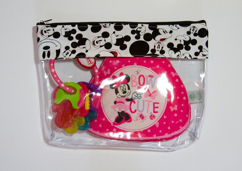 K&ouml;nnte beinhalten: Eine transparente Rei&szlig;verschlusstasche mit einem schwarz-wei&szlig;en Mickey-Mouse-Muster oben. Im Inneren befindet sich ein rosa Babyartikel zum Thema Minnie Mouse und ein farbenfroher Bei&szlig;ring. Der Minnie-Mouse-Artikel tr&auml;gt die Aufschrift "Bow Cute".