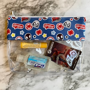 May include: Clear plastic zippered pouch with a blue and white fabric lining featuring a repeating pattern of red and black Spiderman characters, stars, and the text "Let's go Spidey Team!" The pouch contains a yellow lip balm, a clear bottle of hand sanitizer, and a Spiderman comic book.
