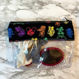 May include: Clear cosmetic bag with a black top featuring colorful cartoon character designs. The bag contains hair accessories and a round, red-rimmed hairbrush. The bag is closed with a black zipper.