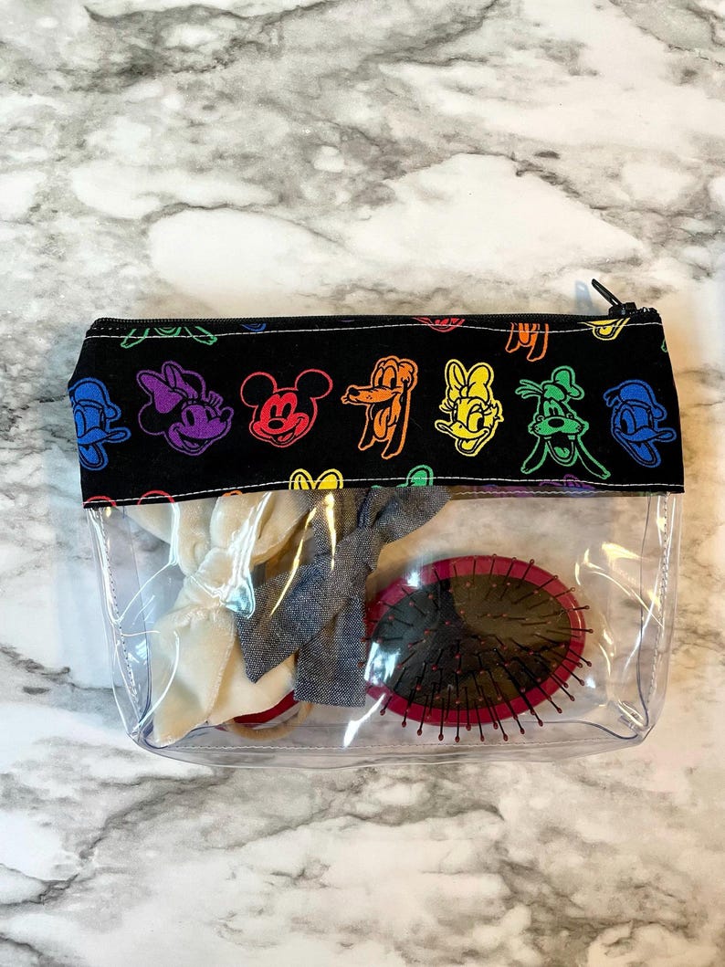May include: Clear cosmetic bag with a black top featuring colorful cartoon character designs. The bag contains hair accessories and a round, red-rimmed hairbrush. The bag is closed with a black zipper.