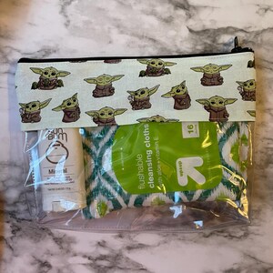 May include: A clear plastic zippered pouch with a white background and a repeating pattern of green baby Yoda characters. The pouch contains a small bottle of sunscreen and a package of flushable cleansing cloths with aloe and vitamin E. The package of cleansing cloths has a green and white geometric pattern and the text "flushable cleansing cloths with aloe + vitamin E" and "up & up" printed on it.