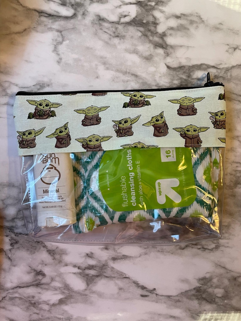 May include: A clear plastic zippered pouch with a white background and a repeating pattern of green baby Yoda characters. The pouch contains a small bottle of sunscreen and a package of flushable cleansing cloths with aloe and vitamin E. The package of cleansing cloths has a green and white geometric pattern and the text "flushable cleansing cloths with aloe + vitamin E" and "up & up" printed on it.