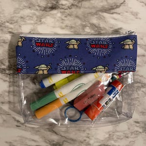 May include: A clear plastic zippered pouch with a blue fabric lining featuring a repeating pattern of a cartoon character and the words "Star Wars". The pouch contains various art supplies, including markers, a glue stick, and an eraser.