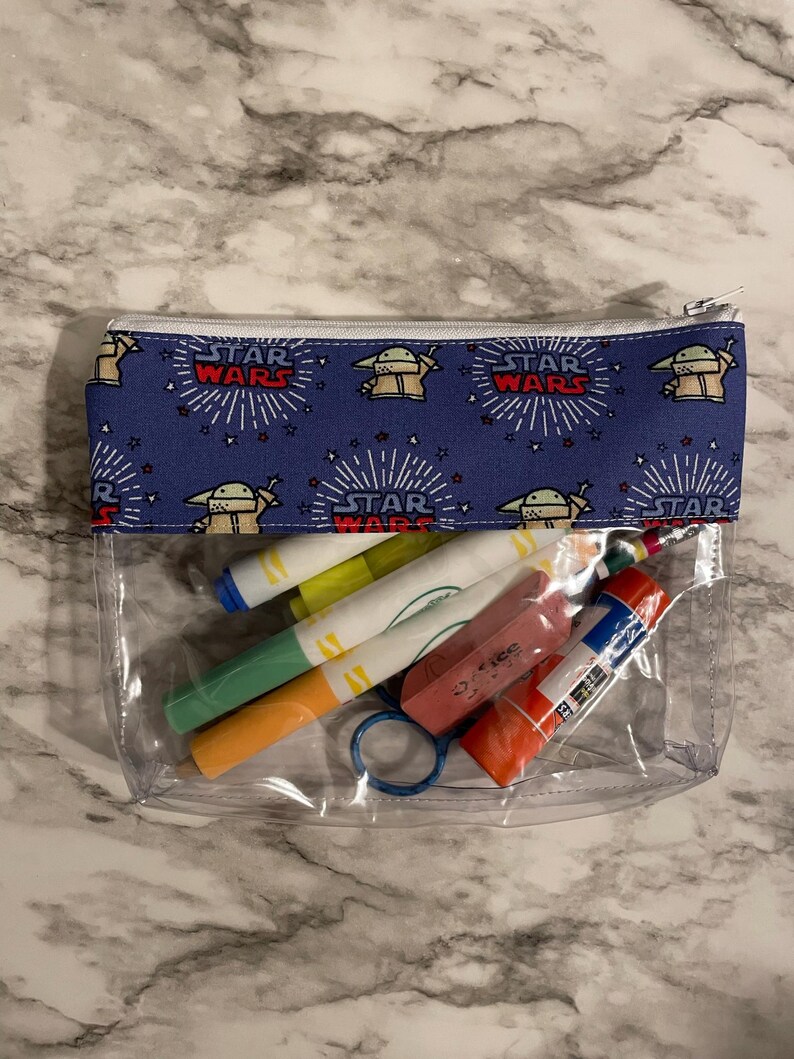 May include: A clear plastic zippered pouch with a blue fabric lining featuring a repeating pattern of a cartoon character and the words "Star Wars". The pouch contains various art supplies, including markers, a glue stick, and an eraser.