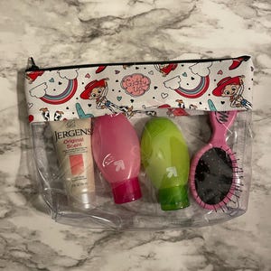 May include: Clear plastic zippered pouch with a white background and a repeating pattern of cartoon cowboys and rainbows. The pouch contains three small bottles of lotion and a pink hairbrush. The text "Giddy Up" is printed on the pouch.