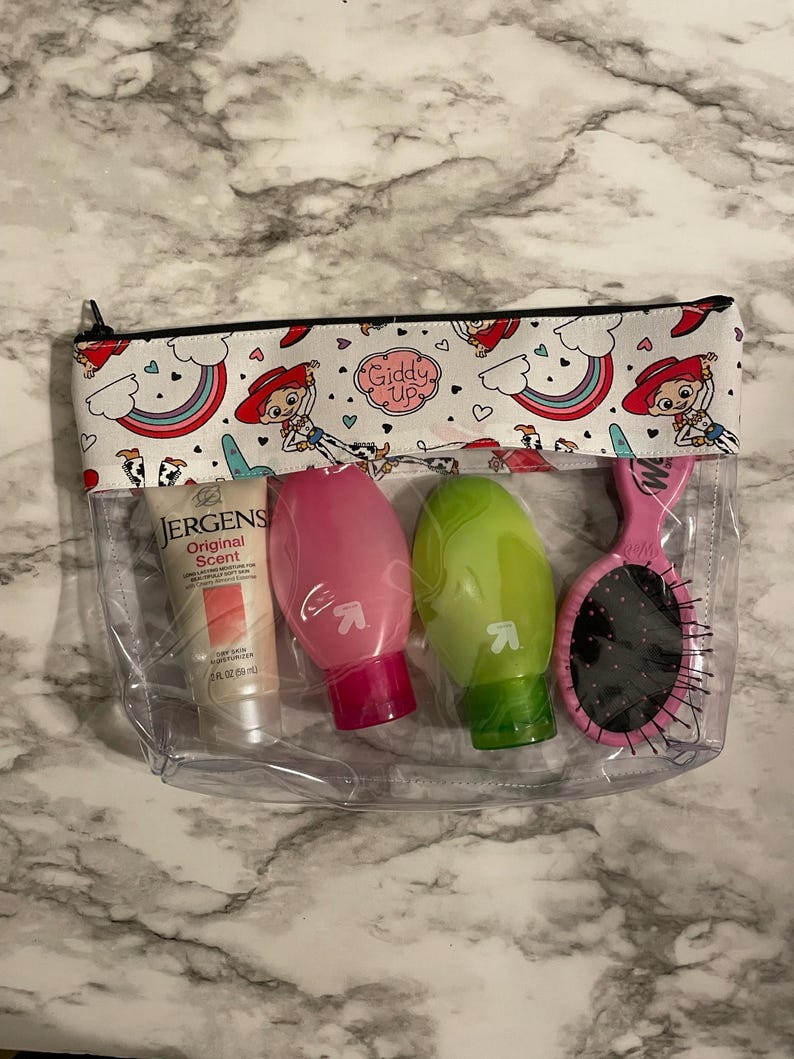 May include: Clear plastic zippered pouch with a white background and a repeating pattern of cartoon cowboys and rainbows. The pouch contains three small bottles of lotion and a pink hairbrush. The text "Giddy Up" is printed on the pouch.