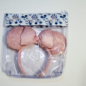 May include: A clear plastic zippered pouch with a white and blue fabric lining featuring a repeating pattern of Mickey Mouse heads. The pouch contains a pair of pink sparkly Minnie Mouse ears.