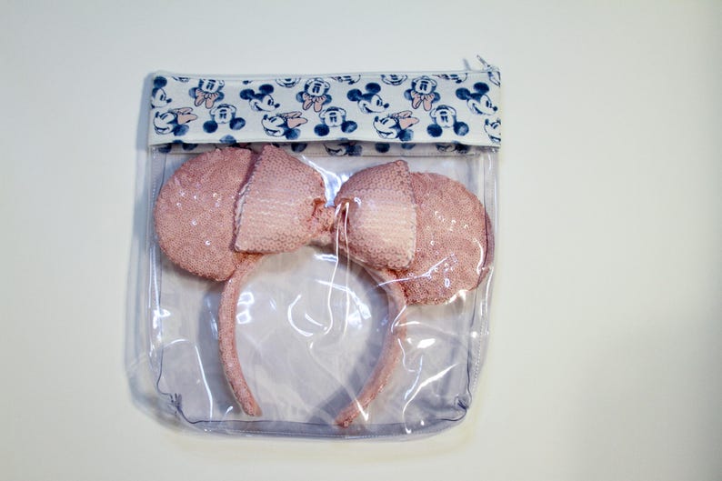 May include: A clear plastic zippered pouch with a white and blue fabric lining featuring a repeating pattern of Mickey Mouse heads. The pouch contains a pair of pink sparkly Minnie Mouse ears.