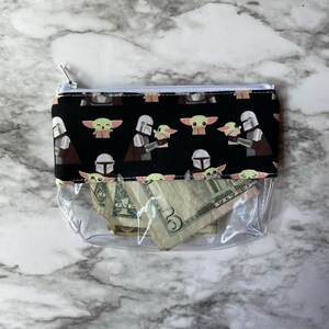 May include: A clear plastic zippered pouch with a black fabric lining. The lining features a repeating pattern of cartoon characters from the Star Wars franchise. The pouch contains several US dollar bills.