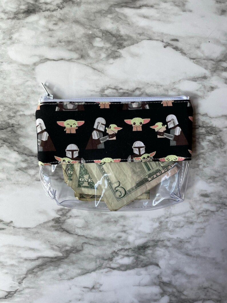 May include: A clear plastic zippered pouch with a black fabric lining. The lining features a repeating pattern of cartoon characters from the Star Wars franchise. The pouch contains several US dollar bills.