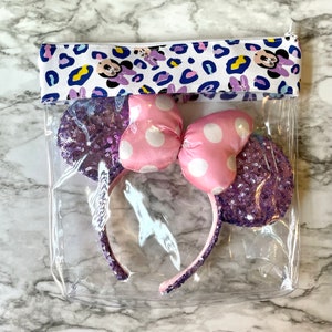 Pode incluir: Uma bolsa transparente com z&iacute;per de pl&aacute;stico com um forro de tecido estampado de leopardo branco e azul. A bolsa cont&eacute;m uma faixa de cabe&ccedil;a Minnie Mouse com lantejoulas roxas e rosa com um la&ccedil;o de bolinhas brancas.