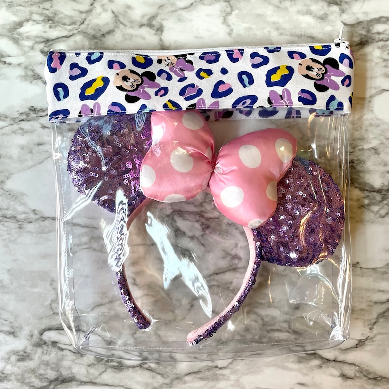 Pode incluir: Uma bolsa transparente com z&iacute;per de pl&aacute;stico com um forro de tecido estampado de leopardo branco e azul. A bolsa cont&eacute;m uma faixa de cabe&ccedil;a Minnie Mouse com lantejoulas roxas e rosa com um la&ccedil;o de bolinhas brancas.
