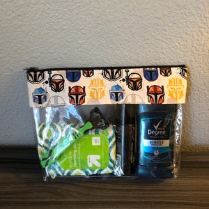 Puede incluir: Una bolsa de pl&aacute;stico transparente con cremallera, con fondo blanco y cremallera negra. La bolsa tiene un patr&oacute;n repetido de cascos Mandalorianos coloridos. La bolsa contiene una maquinilla de afeitar verde, una peque&ntilde;a botella de l&iacute;quido y un desodorante azul y blanco con el texto "Men's Degree Advanced Protection".