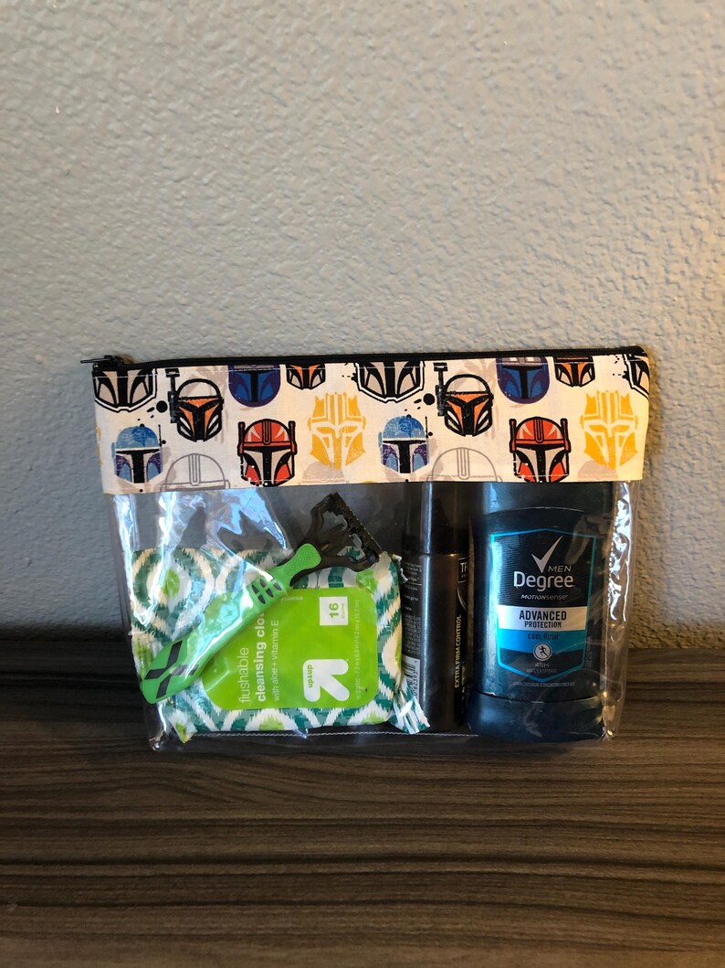 Puede incluir: Una bolsa de pl&aacute;stico transparente con cremallera, con fondo blanco y cremallera negra. La bolsa tiene un patr&oacute;n repetido de cascos Mandalorianos coloridos. La bolsa contiene una maquinilla de afeitar verde, una peque&ntilde;a botella de l&iacute;quido y un desodorante azul y blanco con el texto "Men's Degree Advanced Protection".