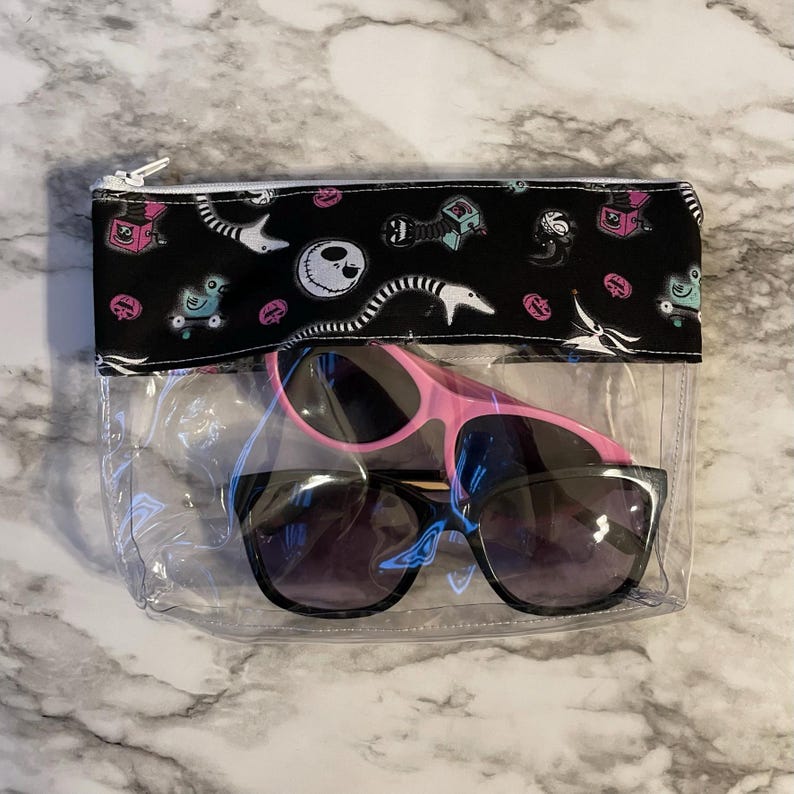 May include: A clear plastic zippered pouch with a black fabric lining featuring a repeating pattern of a character from the Nightmare Before Christmas. The pouch contains two pairs of sunglasses, one pink and one black.