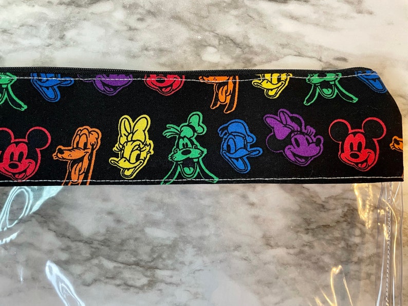 May include: Black pencil case with a zipper closure. The top portion features a repeating pattern of colorful cartoon character faces, including Mickey Mouse, Donald Duck, and Goofy. The bottom half is clear.