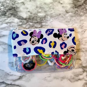 Pode incluir: Uma bolsa transparente com z&iacute;per com um forro de tecido branco que apresenta um padr&atilde;o repetido de cabe&ccedil;as de Minnie Mouse com la&ccedil;os roxos e manchas de leopardo azuis, amarelas e brancas. A bolsa est&aacute; cheia de el&aacute;sticos de cabelo coloridos.