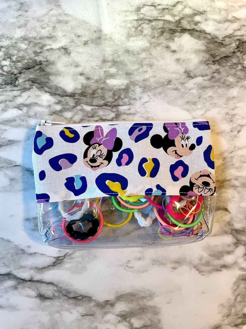 Pode incluir: Uma bolsa transparente com z&iacute;per com um forro de tecido branco que apresenta um padr&atilde;o repetido de cabe&ccedil;as de Minnie Mouse com la&ccedil;os roxos e manchas de leopardo azuis, amarelas e brancas. A bolsa est&aacute; cheia de el&aacute;sticos de cabelo coloridos.