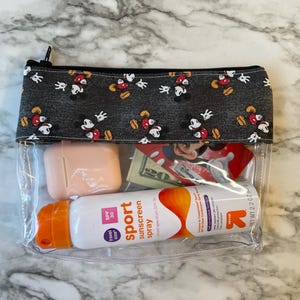 May include: Clear plastic zippered pouch with a black and white Mickey Mouse print fabric lining. The pouch contains a small pink case, a bottle of sunscreen spray labeled "sport sunscreen spray SPF 30", and a few other items.