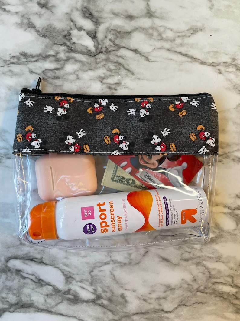 May include: Clear plastic zippered pouch with a black and white Mickey Mouse print fabric lining. The pouch contains a small pink case, a bottle of sunscreen spray labeled "sport sunscreen spray SPF 30", and a few other items.