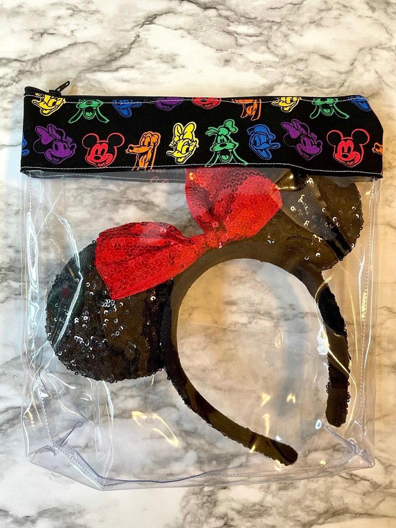 May include: A clear, zippered pouch containing a black sequined headband with a red sequined bow. The top of the pouch is black fabric with colorful cartoon character faces. The pouch is designed to store and protect the headband.