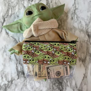 Puede incluir: Una bolsa de pl&aacute;stico transparente con cremallera con un patr&oacute;n marr&oacute;n y verde de un personaje ficticio popular. La bolsa est&aacute; llena de moneda estadounidense, incluyendo un billete de 5 d&oacute;lares, un billete de 10 d&oacute;lares y varios billetes de 1 d&oacute;lar.