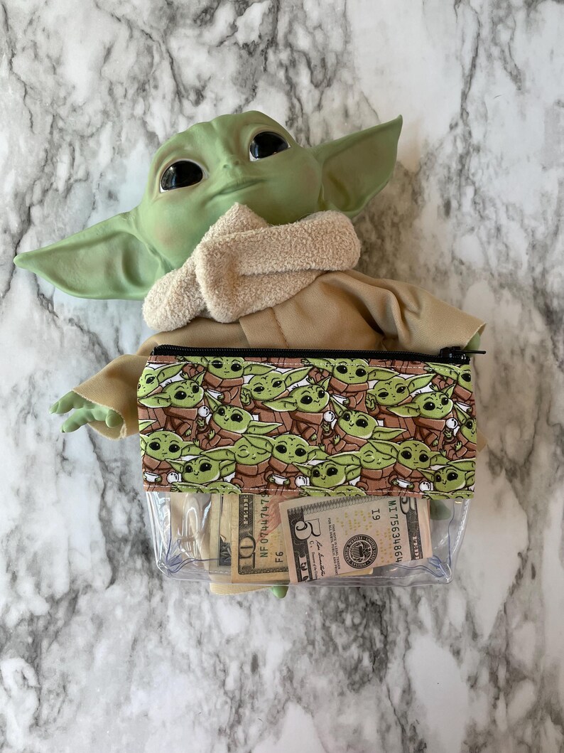 Puede incluir: Una bolsa de pl&aacute;stico transparente con cremallera con un patr&oacute;n marr&oacute;n y verde de un personaje ficticio popular. La bolsa est&aacute; llena de moneda estadounidense, incluyendo un billete de 5 d&oacute;lares, un billete de 10 d&oacute;lares y varios billetes de 1 d&oacute;lar.