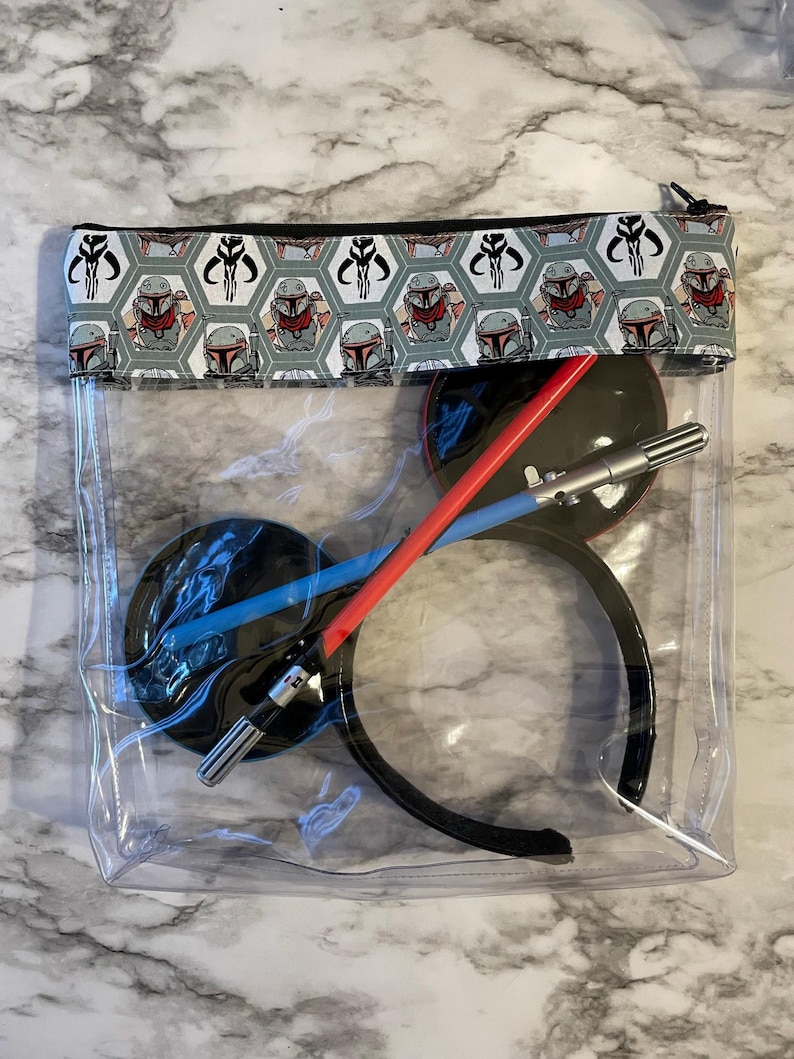 Op de afbeelding: Transparante, doorzichtige ritszak met een zwarte rits en een klep van stof met een herhalend patroon van een groene en grijze Mandalorian helm met een zwart-wit symbool. De tas bevat een zwart hoofdband, een rood-blauw lichtzwaard en een zwart-zilveren lichtzwaard.
