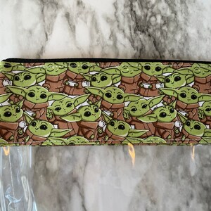 Puede incluir: Una bolsa transparente con cremallera con un forro de tela marr&oacute;n y verde con un patr&oacute;n repetido de un personaje de dibujos animados. El personaje es verde con ojos grandes y sostiene una taza peque&ntilde;a.