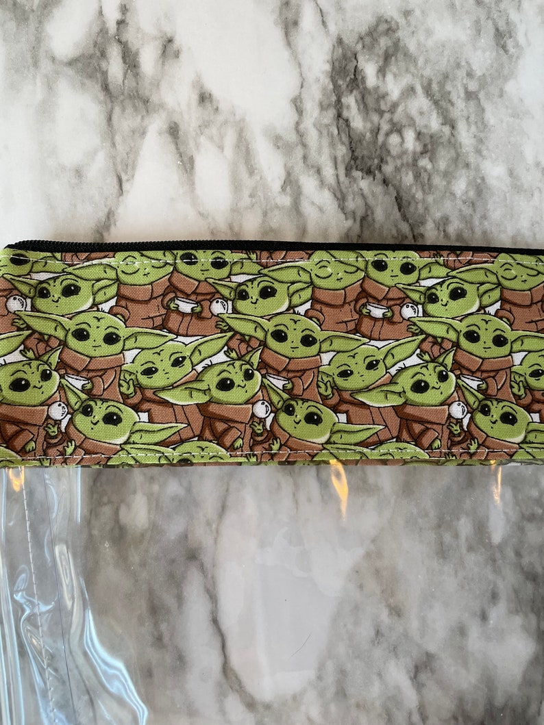 Puede incluir: Una bolsa transparente con cremallera con un forro de tela marr&oacute;n y verde con un patr&oacute;n repetido de un personaje de dibujos animados. El personaje es verde con ojos grandes y sostiene una taza peque&ntilde;a.
