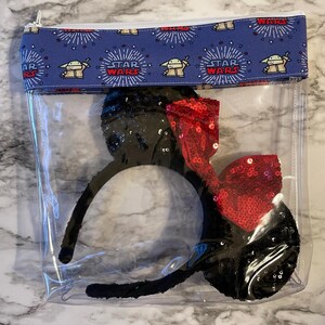 May include: Clear plastic zippered pouch with a blue and white fabric lining featuring a repeating pattern of the Star Wars logo and Baby Yoda. The pouch contains a pair of black sequin Minnie Mouse ears with a red sequin bow.