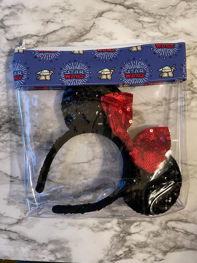 May include: Clear plastic zippered pouch with a blue and white fabric lining featuring a repeating pattern of the Star Wars logo and Baby Yoda. The pouch contains a pair of black sequin Minnie Mouse ears with a red sequin bow.
