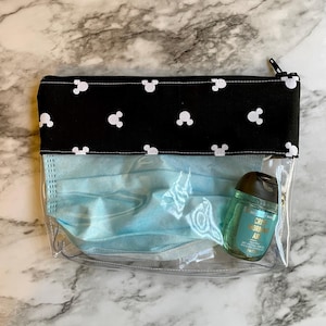 Pu&ograve; includere: Una pochette trasparente con cerniera in plastica con una patta in tessuto nero con un motivo di Topolino bianco. La pochette contiene una maschera facciale blu e un piccolo flacone di disinfettante per le mani etichettato "Crisp Morning Air".