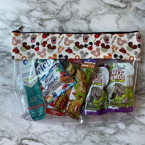 May include: Transparent zipped pouch containing snacks and drinks. The top features a white fabric panel patterned with Mickey Mouse heads, popcorn, and pizza slices. Contents include drink mix, fruit snacks, and fruit bars.