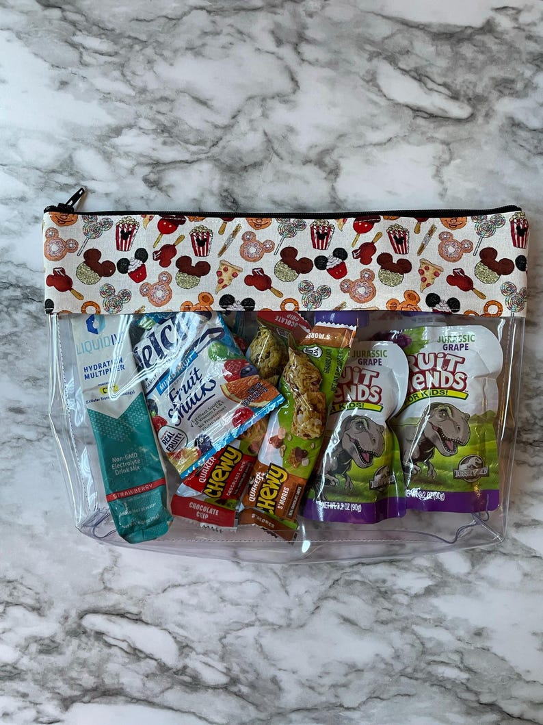May include: Transparent zipped pouch containing snacks and drinks. The top features a white fabric panel patterned with Mickey Mouse heads, popcorn, and pizza slices. Contents include drink mix, fruit snacks, and fruit bars.