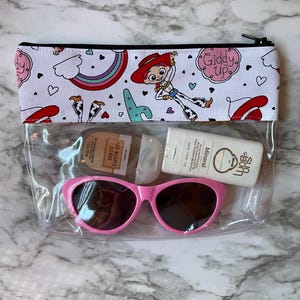 May include: Clear plastic zippered pouch with a white background and a colorful pattern of hearts, rainbows, and a cartoon character wearing a red cowboy hat. The pouch contains a pair of pink sunglasses, a small bottle of sunscreen, and a small bottle of hand sanitizer.