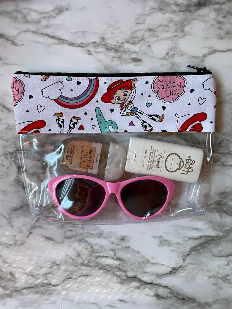 May include: Clear plastic zippered pouch with a white background and a colorful pattern of hearts, rainbows, and a cartoon character wearing a red cowboy hat. The pouch contains a pair of pink sunglasses, a small bottle of sunscreen, and a small bottle of hand sanitizer.