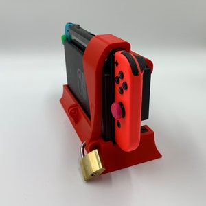 以下が含まれることがあります： ロック付きの赤い3Dプリントスタンド。Nintendo Switch本体とJoy-Conコントローラーを収納するように設計されています。本体は垂直に置かれ、コントローラーはスタンドの側面に取り付けられています。