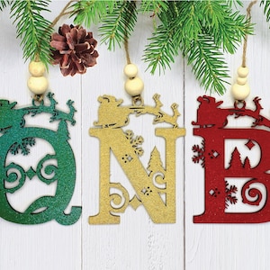 May include: Three wooden Christmas ornaments shaped like the letters Q, N, and B. Each letter is decorated with a festive winter scene and painted in a different color: green, gold, and red.