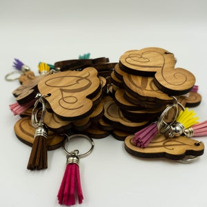 May include: A collection of wooden keychains with a cloud-like shape and a cursive letter design. Each keychain has a silver ring and a colorful tassel in shades of pink, yellow, and brown. The keychains are stacked on a white surface.